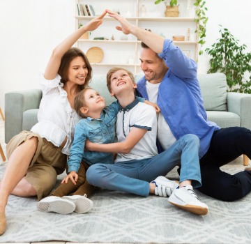 A family of four all white sitting in their living room on the flood on a grey rub with two boys in the middle and mom on the left and dad on the right raising one hand each to make a house over the boys
